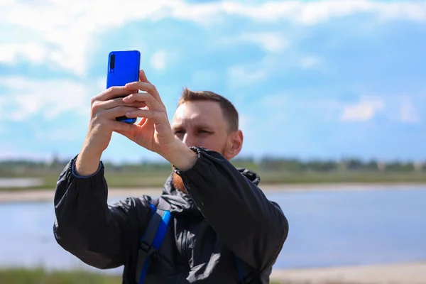 Bearded man in black raincoat on a blue sea landscape background with smartphone in hands takes selfie photography. Portrait of running young hipster guy with cell phone on nature, lifestyle photo.