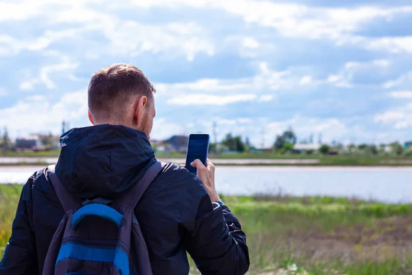 Bearded man in black raincoat on a blue sea landscape background with smartphone in hands takes seascape photography. Portrait of running young hipster guy with cell phone on nature, lifestyle photo.