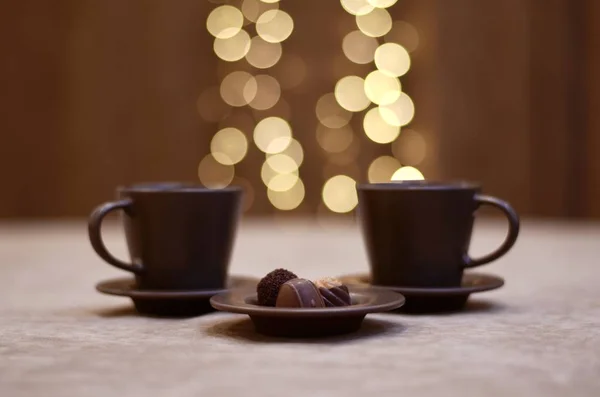 Dos tazas marrones y platillos con luces brillantes sobre ellos y un plato de chocolates espolvoreado con cacao . — Foto de Stock