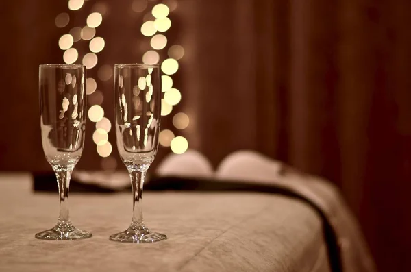 Dois copos de vinho brilhantes mais perto na cama e silhuetas borradas de roupas no quarto com fogos bokeh . — Fotografia de Stock