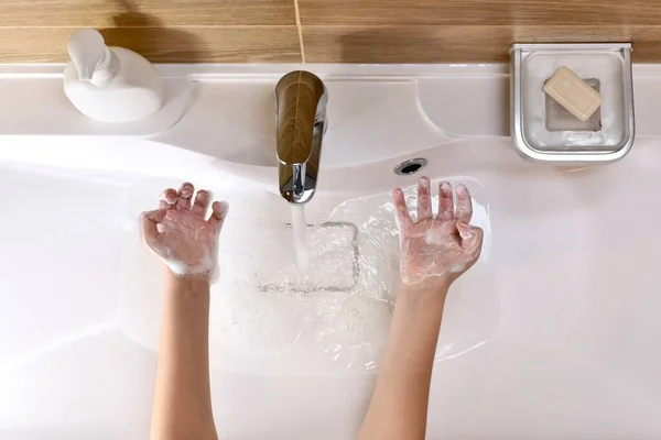 El niño muestra las palmas enrolladas por las manos llenas de espuma jabonosa, con los dedos doblados, por encima del fregadero con agua que vierte . —  Fotos de Stock