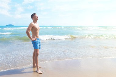 Mavi denizdeki kumsalda yüzme şortu giymiş yakışıklı, zayıf, atletik bir adamın tam boy portresi. Genç adam tropikal tatilden, yaz güneşli havadan, güneşlenmekten, yüzmekten hoşlanıyor..