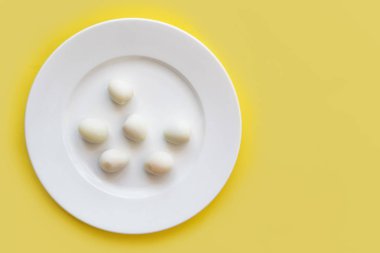 Sarı arka planda beyaz tabakta soyulmuş organik, küçük, haşlanmış bıldırcın yumurtaları. Üst görünüm, boşluğu kopyala. Sağlıklı protein taze doğal besin. Doğal gurme yemeği. Küçük kuş yumurtaları.