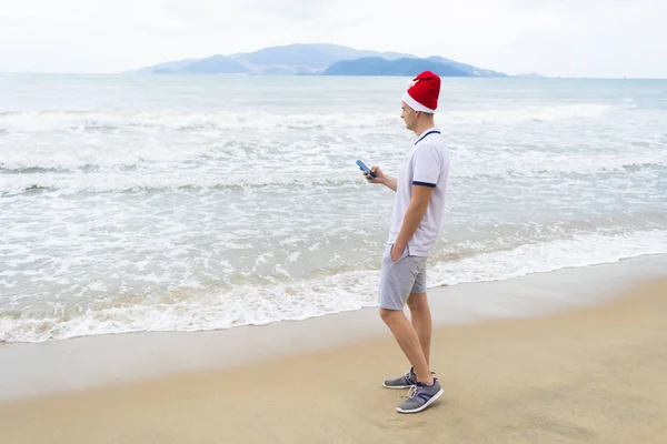 Handsome sad guy on the beach, sea, sand standing near ocean in Santa Claus red hat, looking at cell mobile smart phone, typing message. Merry Christmas, lonely New Year alone far from home. Vacation.