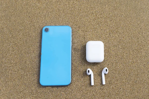 New modern blue phone, smartphone with earphones, wireless case on clear yellow sand, beach, sea. Top view. Travel, vacation with gadgets, devices.