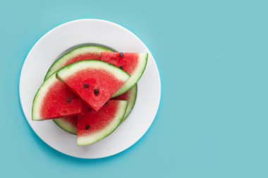 Mavi arka planda beyaz tabakta olgun kırmızı karpuz dilimleri. Üst manzara, düz yatış. Boşluğu kopyala, metin için yer aç. Vejetaryenler için sağlıklı çiğ yiyecekler, vejetaryenler, yaz tatili konsepti. Meyve kes