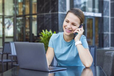 Mutlu neşeli kız, genç meşgul meşgul güzel kadın gülümseyen, sohbet eden, cep telefonuyla konuşan akıllı telefon, dizüstü bilgisayarına bakan, dışarıda kafede bilgisayar kullanan. Teknoloji ve insanlar kavramı
