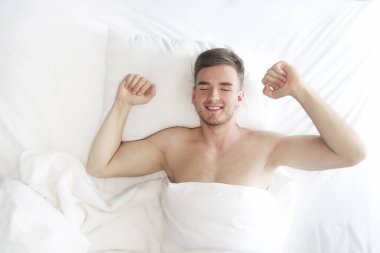 Mutlu, neşeli, genç, yakışıklı bir adam yatakta yatıyor, uyuyor, uyanıyor, esniyor, günün tadını çıkarıyor, günaydın, gülümsüyor. Beyaz yatak üzerinde sağlıklı bir uyku. Rahatlama, gözler kapalı.