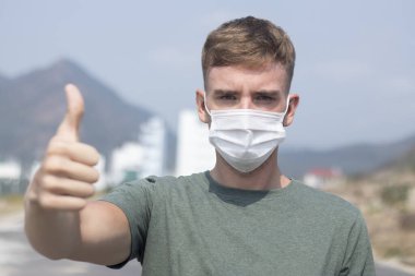 Ciddi Avrupalı bir adam, koruyucu steril tıbbi maskeli genç yakışıklı bir adam kameraya bakıyor baş parmağını kaldırıyor, jest gibi. Hava kirliliği, virüs, Çin pandemik koronavirüsü koruma konsepti
