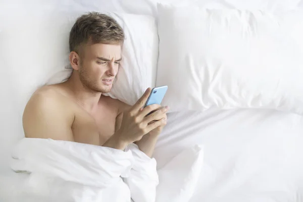 Handsome negative disgusted sad man, young upset angry tired guy lying in bed on pillow with cell mobile, using smart phone, get bad news, argue with wrinkled displeased face before sleep, sleeping