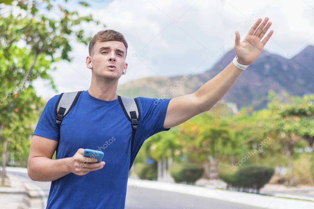 Handsome guy, young man in hurry late, trying to stop, catch a taxi car or cab. Boy standing near road with backpack and cell mobile smart phone, raise, waving his hand, holding arm up. Hitchhiker