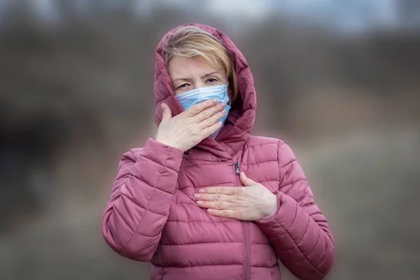 Senior sick ill serious woman in medical sterile mask on her face coughing, holding hand on her chest, lungs, looking at camera. Pandemic, virus, covid-19 concept. Coronavirus symptoms — Stock Photo, Image