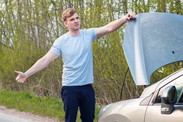 Gefrustreerd trieste man bestuurder met een kapotte auto, motor, motor met open motorkap. Verkeersongeval, ongeval. Jonge wanhopige man kreeg een verkeersongeluk, geïrriteerde man liftend en vroeg om hulp. — Stockfoto