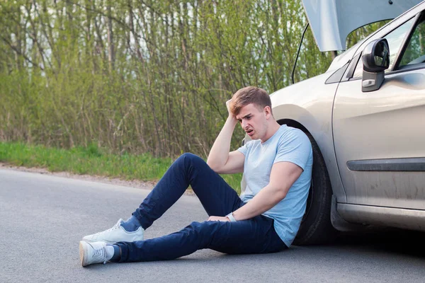 Frustrado triste conductor tipo molesto está sentado cerca de un coche roto después de accidente de tráfico, accidente. Joven sorprendido desesperado hombre asustado entró en accidente de tráfico, sentado en asfalto sosteniendo la cabeza con la mano —  Fotos de Stock