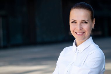 Güzel, mutlu, pozitif bir genç kadın, başarılı, neşeli, beyaz tişörtlü iş kadını açık hava iş merkezinde kameraya bakıp gülümsüyor. Avrupalı kadın portresi, kopyalama alanı