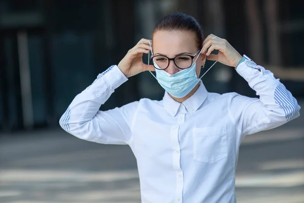 Beautiful business woman, young girl putting on medical protective mask on her face, in white shirt in glasses outdoors, healthcare at job, work, office. Coronavirus, virus, epidemic, covid-19 concept — Stock Photo, Image