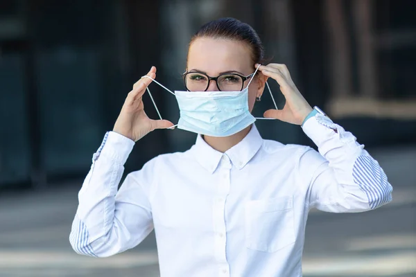 Beautiful business woman, young girl putting on medical protective mask on her face, in white shirt in glasses outdoors, healthcare at job, work, office. Coronavirus, virus, epidemic, covid-19 concept — Stock Photo, Image
