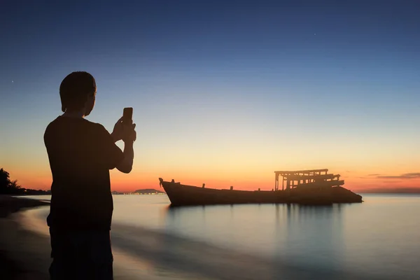 Hombre silueta utilizar el teléfono inteligente tomar una foto en el mar con sunr —  Fotos de Stock