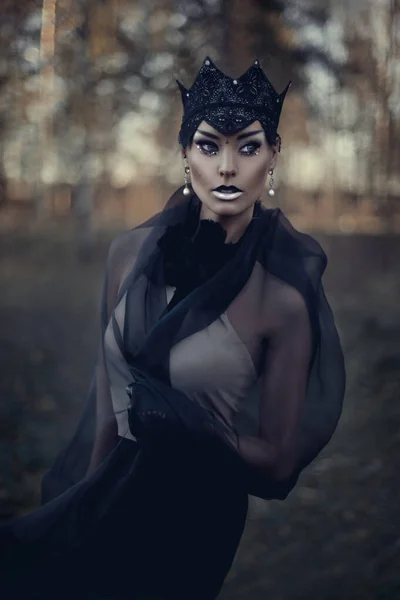 Gothic beauty, the dark queen. Art Photography. A beautiful gothic princess with make up and black crown and a black long dress walks in a misty fairytale autumn forest. The costume of the dark queen.