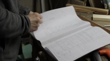 Erkek marangoz, ahşap bina planları için defteri kontrol ediyor, bazılarını gösteriyor ve detayları anlatıyor.