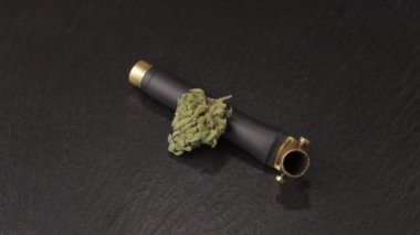 Brass weed smoking pipe with sativa and indica weed bud on turning slate board table. Black background