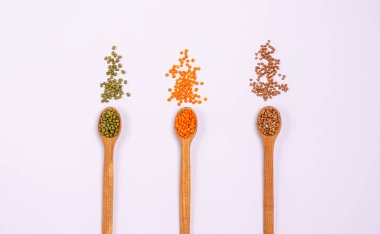 Tahta kaşıklardaki vejetaryenler için çeşitli kuru tahıllar: yeşil ve turuncu mercimek, beyaz arka planda kahverengi karabuğday. Protein ve karbonhidrat açısından sağlıklı gıdaları kopyalayın. Yukarıdan görüntüle.