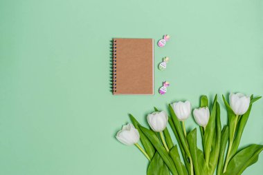 Bir buket beyaz lale, kraft kağıdından bir not defteri ve yeşil arka planda dekoratif mandallar, kopyalama alanı, düz yatış. Mesaj için yer. Tatil konsepti, bahar. Yukarıdan görüntüle.