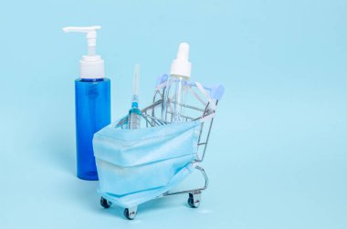 Koruyucu tıbbi maskesi, şırıngası, antiseptiği ve mavi arka planda sabunu olan dekoratif küçük alışveriş arabası..