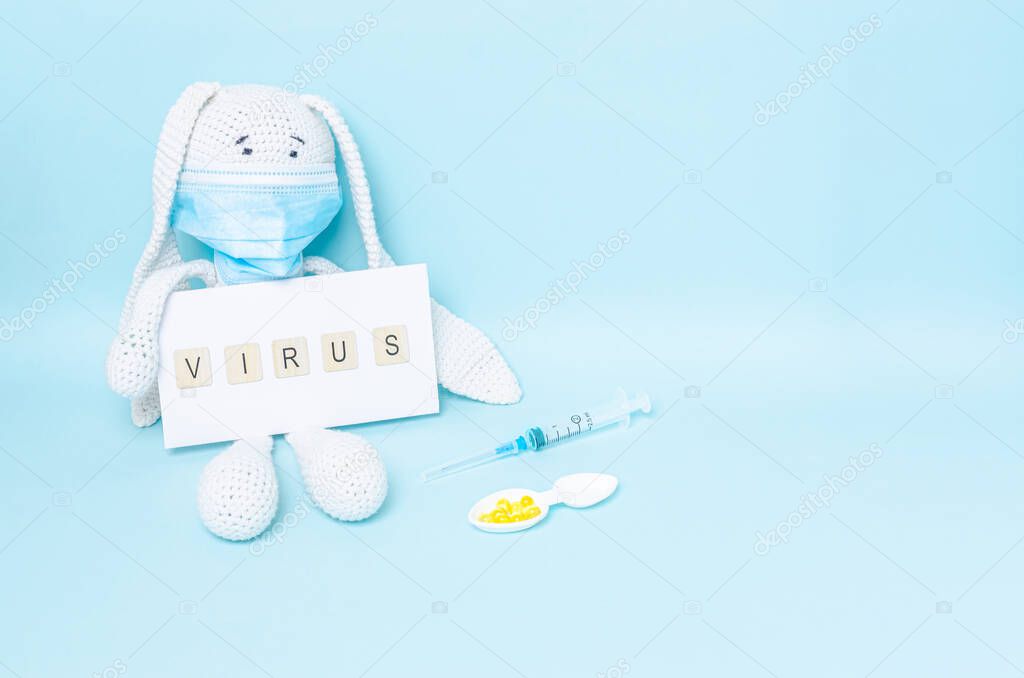 White soft toy with a protective medical mask and the inscription virus, yellow pills in a spoon and a syringe on a blue background