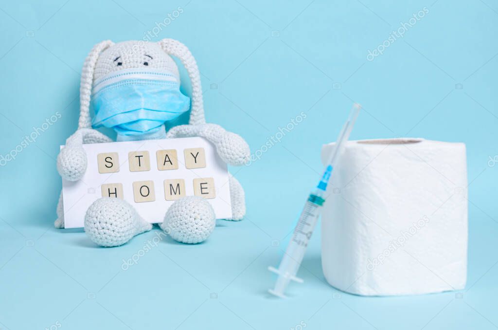 Toilet paper and syringe, children's soft toy bunny in a protective medical mask holds the inscription stay home on a blue background. Coronovirus, Covid-19, Quarantine, Pandemic. Close-up. Copy space