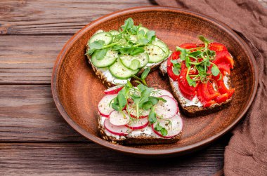 Kil tabakta krem peynirli ve sebzeli çeşitli mini sandviçler. Peynirli sandviç, salatalık, turp, tatlı biber, ahşap zemin üzerinde mikroyeşil. Düz konum, kopyalama alanı. Yakın plan.