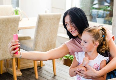 Gülümseyen bir kız akıllı telefondan tatlı bir kızla selfie çekiyor, mutlu bir genç anne gülüyor, küçük kızıyla fotoğraf çekiyor, telefonla oynuyor, cep telefonuyla aile videosu kaydediyor.