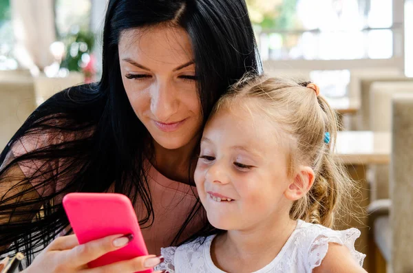 Gülümseyen bir kız akıllı telefondan tatlı bir kızla selfie çekiyor, mutlu bir genç anne gülüyor, küçük kızıyla fotoğraf çekiyor, telefonla oynuyor, cep telefonuyla aile videosu kaydediyor.