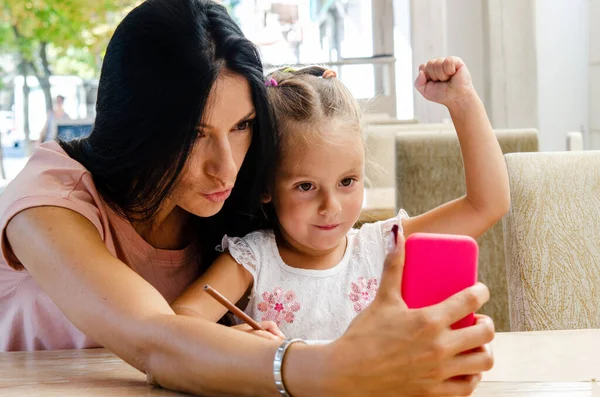 Gülümseyen bir kız akıllı telefondan tatlı bir kızla selfie çekiyor, mutlu bir genç anne gülüyor, küçük kızıyla fotoğraf çekiyor, telefonla oynuyor, cep telefonuyla aile videosu kaydediyor.