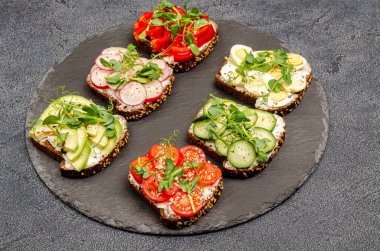 Siyah bir tabakta krem peynirli ve sebzeli mini sandviçler, siyah arka plan. Peynirli sandviç, turp, avkado, yumurta, salatalık, tatlı biber, domates, mikroyeşil, üst manzara