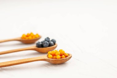 Beyaz arka planda olgun yaban mersini ve ahşap kaşıklarda deniz dikenli üzümler. Boşluğu, düzlüğü, metin için yeri kopyala. Doğal tatlı, sağlıklı atıştırmalık. Yakın plan..