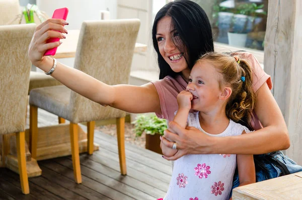 A smiling girl takes a selfie with a cute girl on a smartphone, a happy young mother laughs,takes a photo with her little daughter,playing fun with the phone,recording family video on her mobile phone