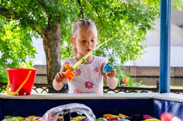 Beyaz elbiseli küçük bir kız utangaç bir şekilde bir oyuncak yakalıyor, çocuk havuzunda balık tutuyor. Havuzda çocuk oyuncakları var. Oyuncak olta kamışı. Çocuklar için balık tutmak, çocuk havuzunda balık tutmak.