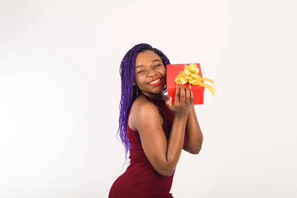 Uma menina bonita preta em um vestido festivo está feliz em receber um presente para o feriado. Uma linda garota afro-americana segura um grande presente vermelho em suas mãos, sorri e olha para a câmera. Isolado em — Fotografia de Stock