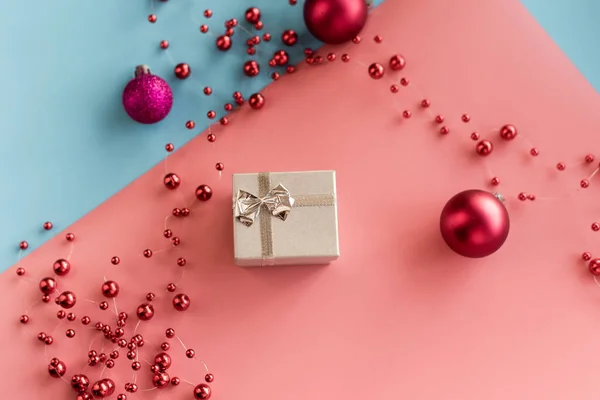 Piccolo regalo d'argento su sfondo rosa e blu pastello con palline di Natale rosse e perline. Il concetto di dare regali, sorprese, Natale, Capodanno — Foto Stock