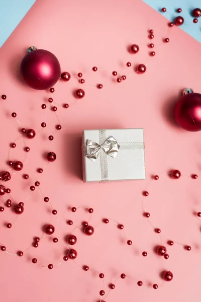 Sølvfarget liten gave på rosa og blå pastellbakgrunn med røde juleballer og perler. Begrepet "å gi gaver, overraskelser, jul, nytt år" – stockfoto