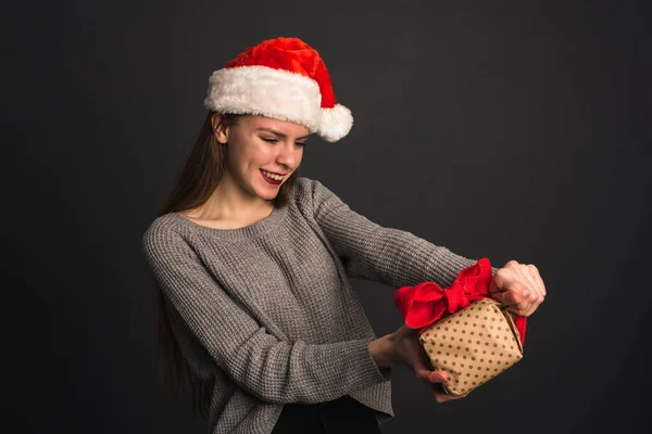 Una hermosa chica con un sombrero de Santa Claus sobre un fondo gris oscuro abre un regalo de Navidad y rompe el embalaje de regalo de Kraft. La modelo rubia rompe el embalaje artesanal de un regalo de Navidad. El cono —  Fotos de Stock