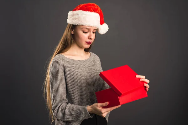 Un modelo en un sombrero de Santa Claus está decepcionado con el regalo. La chica abre el regalo rojo de Navidad y muestra la emoción de sorpresa y decepción —  Fotos de Stock