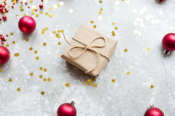 Een klein cadeautje versierd met kraftpapier en touw op een grijze achtergrond met goudconfetti, rode kerstballen en een slinger. Het concept van winkelen, cadeaus kopen, verrassingen geven en ontvangen, nieuwe — Stockfoto