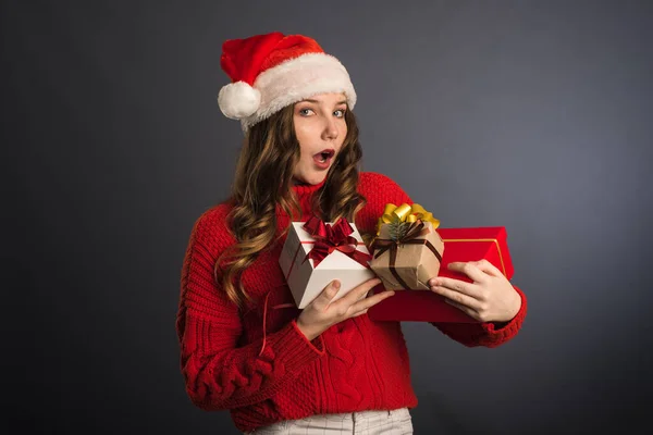 Chica sorprendida con un montón de regalos en sus manos. Una hermosa chica con una cara expresando la emoción de wow, en un suéter rojo y un sombrero de Navidad Santa Claus. El concepto de la Navidad y el nuevo yo —  Fotos de Stock