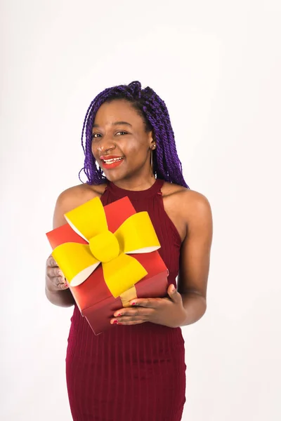Egy sötét bőrű, gyönyörű, afrikai copfos modell egy ajándékkal a kezében húzza a kezét. Fekete aranyos lány ad egy nagy piros ajándékot. A meglepetés, a nyaralás és a vásárlás fogalma. Sablon — Stock Fotó