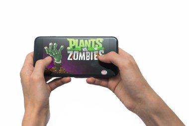 Samara, Russia -07, 29, 2019: A young guy plays a plants vs. zombies game on Iphone 8 Plus. Teenage boy holding a phone in his hands with a game plants vs zombies mobile on a white background
