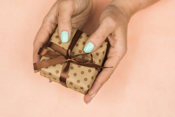 Mãos de uma menina com uma manicure pastel azul segurando um presente de artesanato com um arco em um fundo rosa. Conceito de férias, presentes, surpresas, aniversário, ano novo e Natal — Fotografia de Stock