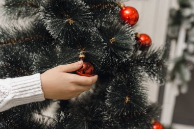 Beyaz örgü süveter giyen bir el yapay bir Noel ağacının dalına kırmızı bir Noel topu asar. Noel ağacını oyuncaklarla süslemek için yakın plan.