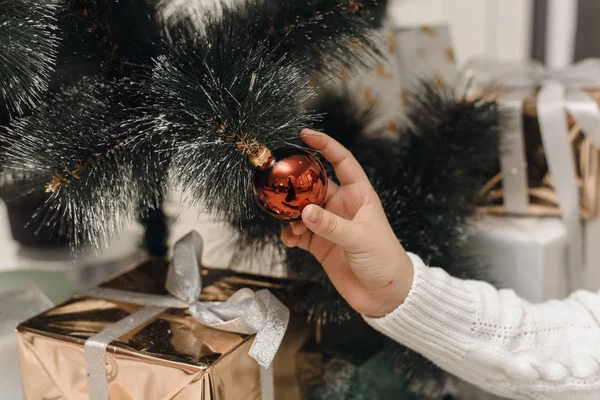 Eine Hand in einem weißen Strickpullover hängt eine rote Weihnachtskugel am Ast eines künstlichen Weihnachtsbaums. Der Weihnachtsbaum aus nächster Nähe mit Spielzeug dekorieren. — Stockfoto